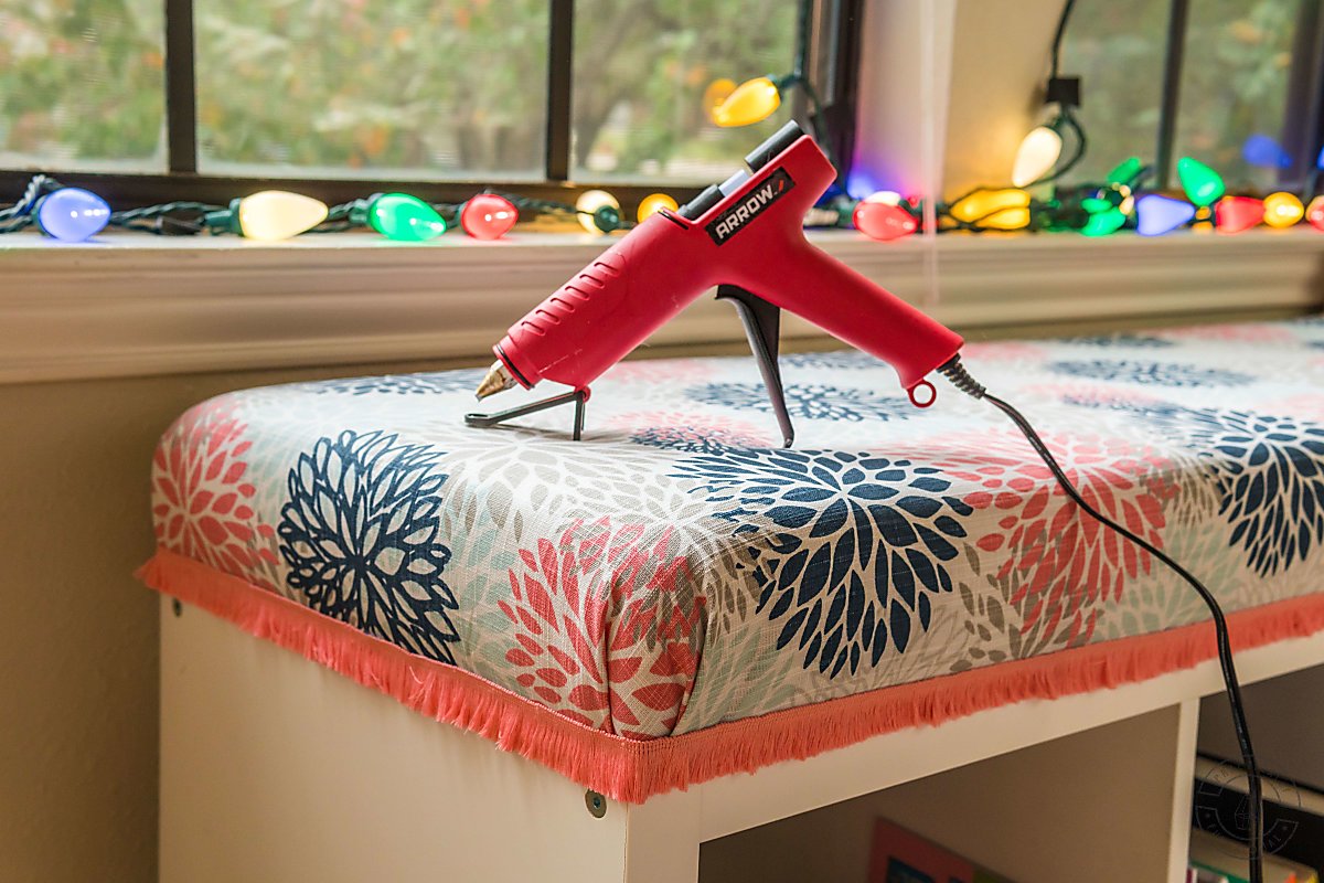 hot glue gun on top of diy bench seat cushion with pink fringe trim around the bottom of the cushion